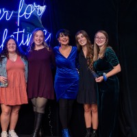 Group of 5 girls and President Mantella stand in front of Laker for a Lifetime neon sign with someone's hand in the corner taking the picture